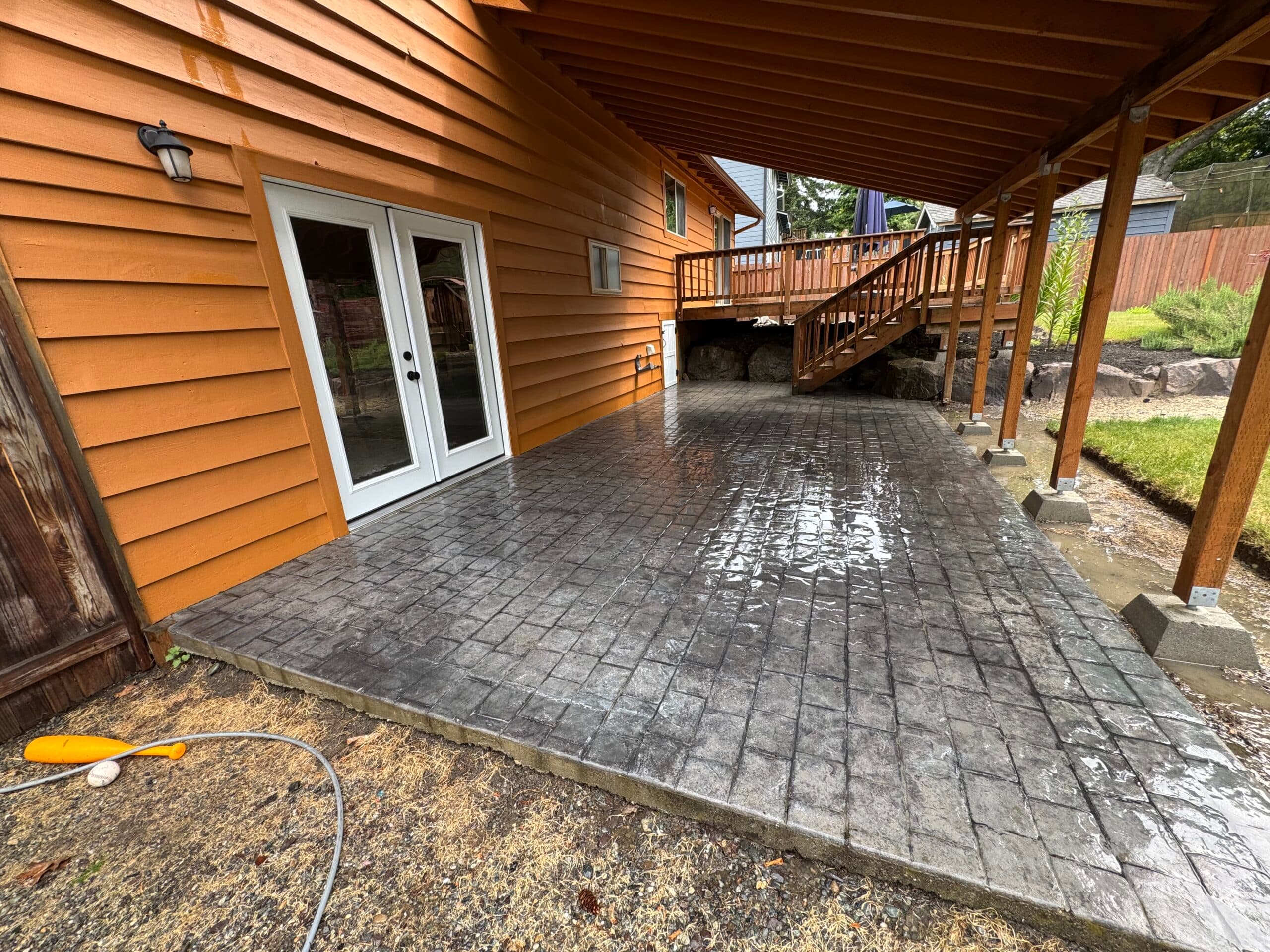 Freshly washed dark stamped-concrete patio glistening wet beside a cedar-sided house